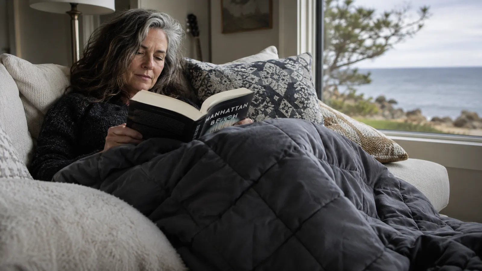 A neatly made bed near a coastal window at dawn with a dark grey weighted blanket folded back on white linen, soft morning light coming in