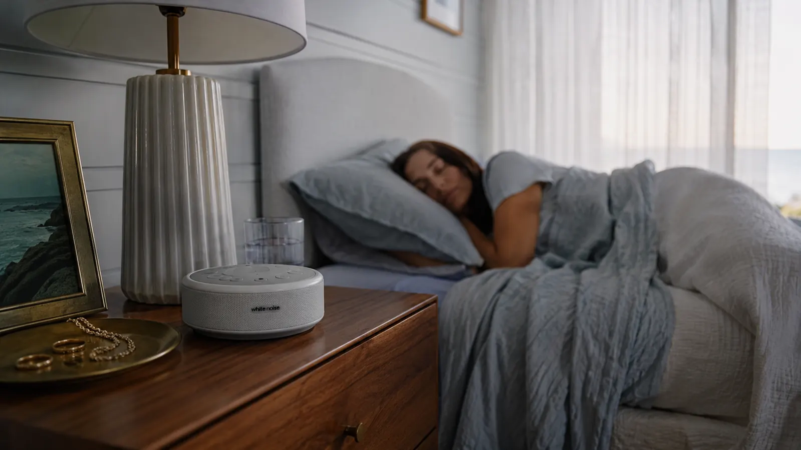 Small white noise machine glowing softly on a wooden nightstand beside a glass of water and a sleeping woman in dim coastal bedroom light